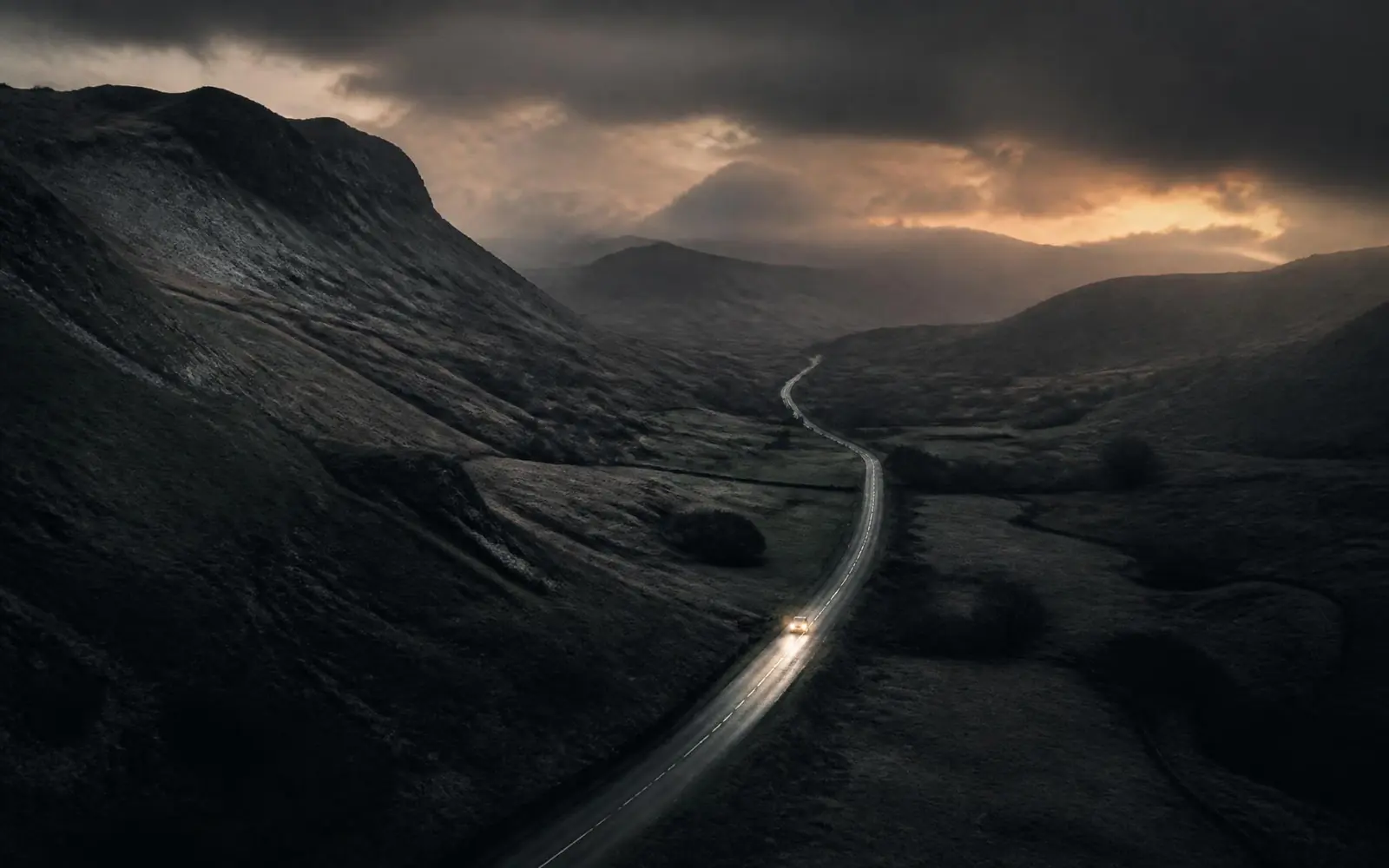 A road cutting through dramatic fells at dusk, a single vehicle heading out